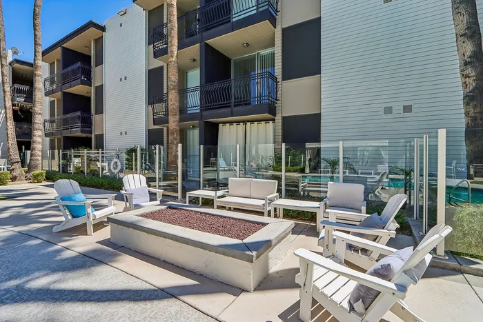 Charming outdoor patio area with comfortable seating and a fire pit, perfect for gatherings. Modern apartment complex with glass balcony railings and a pool view.