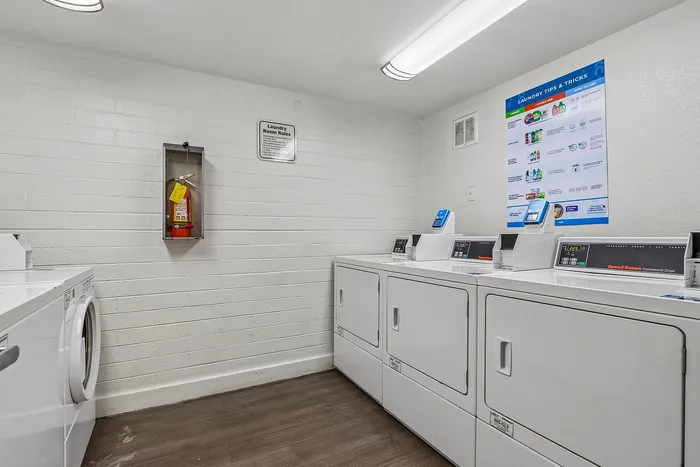 Modern laundry room featuring commercial Speed Queen dryers, vibrant informational posters, and sleek white appliances against a chic brick wall backdrop.