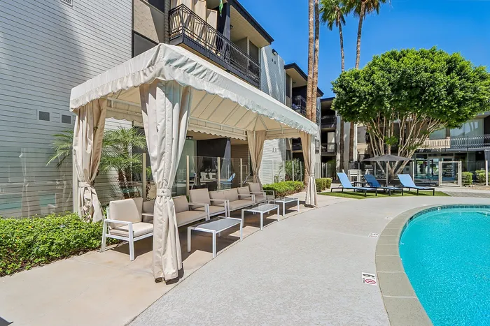 Inviting outdoor lounge area with a stylish cabana and comfortable seating by a sparkling blue pool, surrounded by lush greenery and modern apartment buildings.