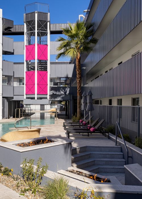 Modern apartment courtyard featuring a pristine pool, stylish sun loungers, vibrant pink accents, and a cozy fire pit surrounded by lush greenery.