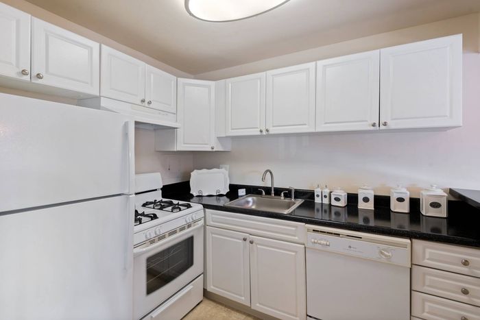 Laurel, MD, Apartments Near Tanger Outlets - SouthRidge Apartments & Townhomes - Kitchen With White Cabinetry And Appliances, Sleek Black Countertops, A Stainless Steel Sink, And Ceramic Canisters.
