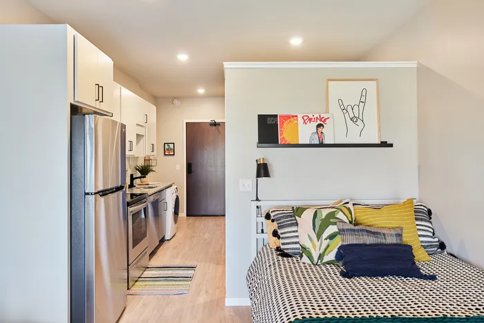 Pet-Friendly Apartments in Minneapolis, MN - B-Side - Cozy Studio Kitchen With Bed, Stainless Steel Appliances, White Oak Flooring, Recessed Lighting, and Entry Hallway View.