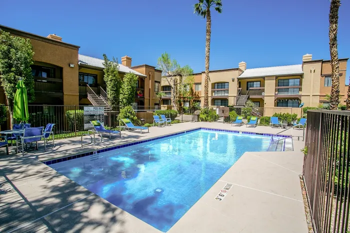 North Las Vegas, NV, Apartments With In-Unit Washer and Dryer - Desert Ridge Apartments - Outdoor Swimming Pool With Lounge Chairs, Seating With Umbrella, Trees, Metal Fence, and Apartment Buildings.