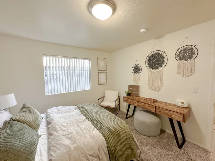 Pet-Friendly Apartments in West Las Vegas, NV - Portola Del Sol Apartment Homes - Bedroom With Green Throw Blanket, Large Window With Vertical Blinds, Accent Chair, And Wooden Desk.