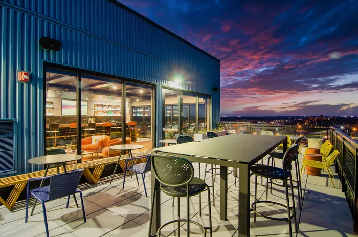 Apartments in Minneapolis, MN, With In-Unit Washer and Dryer - B-Side - Rooftop Lounge With Community Tables, Bar Seating, Outdoor Chairs, and Glass Doors To Resident Lounge With City Views at Dusk.