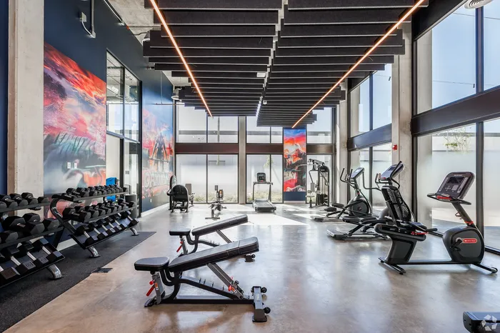 Apartments in West Oakland, CA, with In-Unit Washer and Dryer - Prescott Station - A Modern Fitness Center Features Weight Benches, Dumbbell Racks, Cardio Machines, And Floor-To-Ceiling Windows.