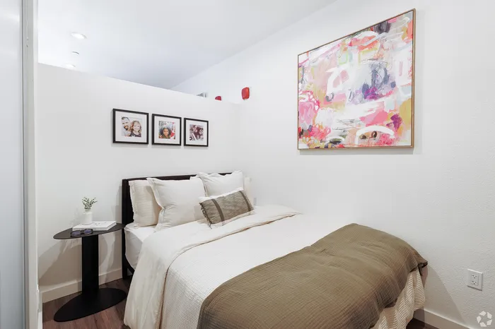 Pet-Friendly Apartments in West Oakland, CA - Prescott Station - Bedroom Features A Neatly Made Bed With White Bedding, A Brown Throw Blanket, A Side Table, Framed Photos, And Wood-Style Floors.