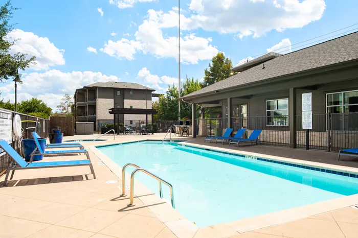 Dog-Friendly Apartments in North Austin, TX - Lotus Village Apartments - An Outdoor Swimming Pool With Blue Lounge Chairs, Poolside Deck, And Clubhouse Building.