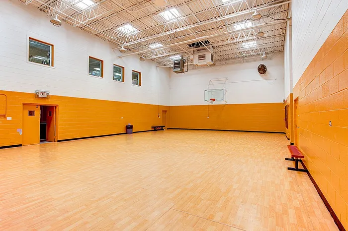 Justice, IL, Apartments Near Bridgeview - Willow Hill Apartments - Indoor Basketball Court With Polished Wood Floor, High Ceiling Lights, Orange Accent Walls, and Hoop Backboard.