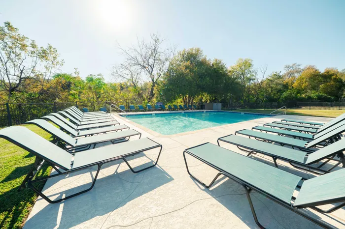 Two-Bedroom Apartments in Austin, TX - Agave Falls - Outdoor Swimming Pool Surrounded by Lounge Chairs, Lawn, Metal Fence, Lush Trees, and Light-Colored Concrete Deck.