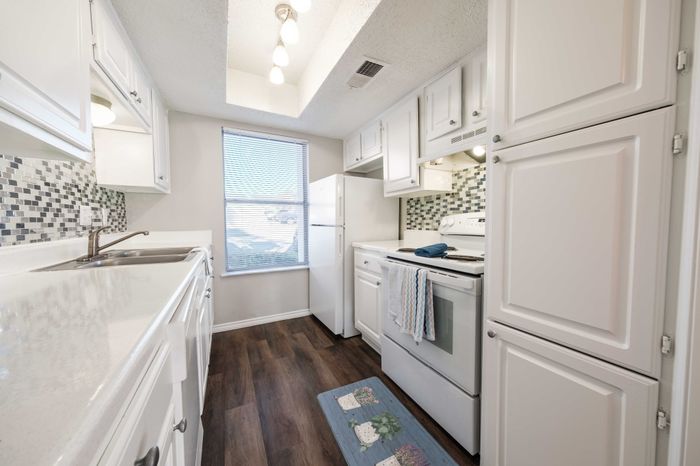 Dog-Friendly Apartments in Austin, TX - Agave Falls - A White Kitchen with Wood Flooring, White Appliances, Double Sink, Light Fixtures, Area Rug, and Window with Blinds.