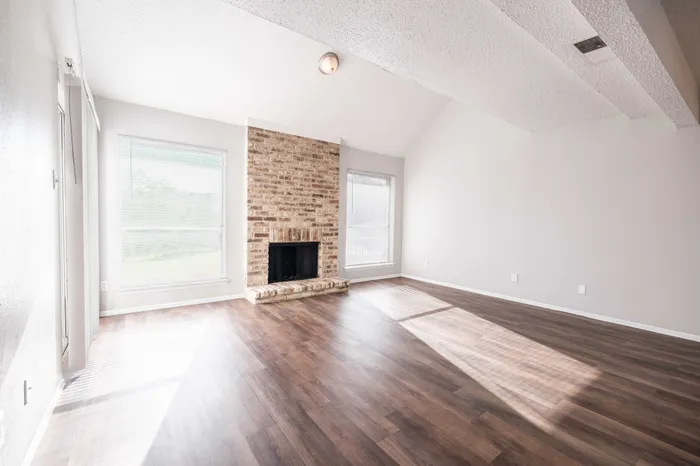 Austin, TX, Pet-Friendly Apartments in Austin, TX - Agave Falls - Spacious Living Area with Brick Fireplace, Vaulted Ceiling, Large Windows with Blinds, and Dark Wood-Style Flooring in Bright Open Layout.