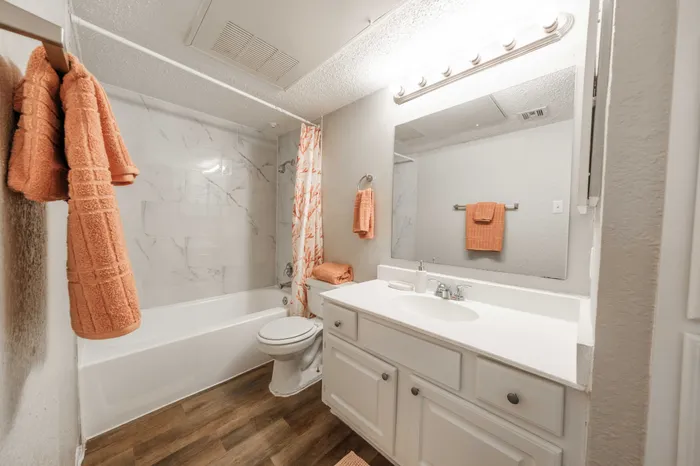 Pet-Friendly Apartments in Austin, TX - Agave Falls - White Bathroom with Bathtub, Tile Surround, Wood Flooring, Single-Sink Vanity, Mirror, and Light Fixtures.