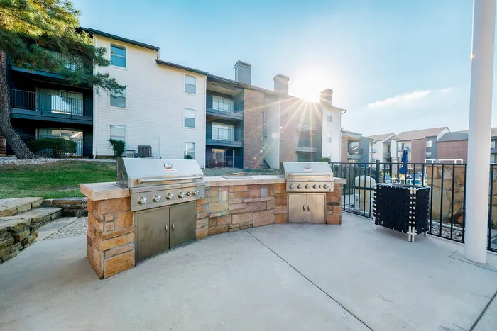 Dog-Friendly Apartments in Irving, TX - Bridgeport Apartment Homes - Outdoor Grilling Station with Built-in Stainless Steel Gas Grills, Stone Counter, Black Fence, and Apartment Buildings View.