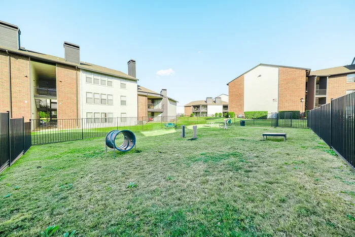 Irving, TX, Pet-Friendly Apartments - Bridgeport Apartment Homes - Fenced-in Bark Park with Open Grass Area, Agility Tunnel and Obstacles, Benches, and Surrounding Apartment Buildings on a Clear Day.