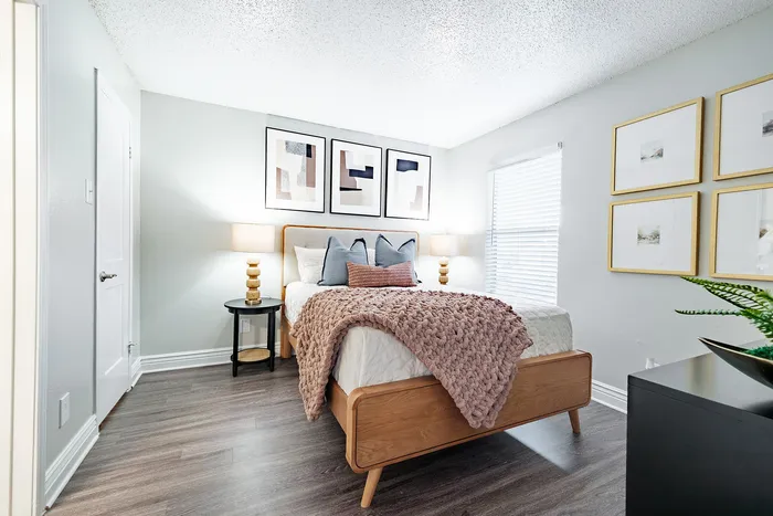 Irving, TX, Apartments Near Las Colinas - Bridgeport Apartment Homes - Bedroom with Wood Bed Frame, Plush Bedding, Nightstands, Soft Lamp Lighting, Wood Flooring, and Window with Blinds.