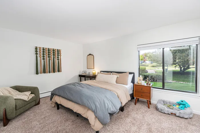 1-Bedroom Apartments in Palatine, IL - The Clayson - A Bedroom With Carpeted Flooring, Nightstands, 2-Seater Couch, Wall Art, And A Large Window.