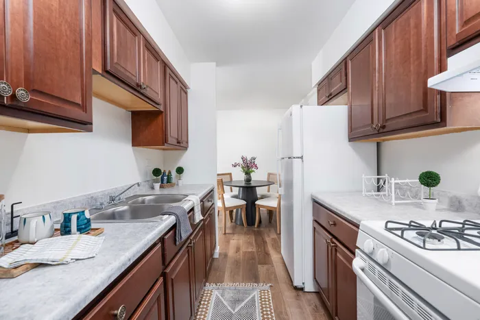 Pet-Friendly Apartments in West Chicago, IL - Timber Lake Apartment Homes - A Galley Kitchen With Dark Wood Cabinets, White Appliances, Gray Countertops, Double Sink, And Gas Stove.