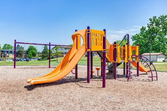 Justice, IL, Pet-Friendly Apartments - Willow Hill Apartments - Community Playground With Orange Slides, Climbing Bars, Swing Set, and Open Green Space Under Blue Sky.
