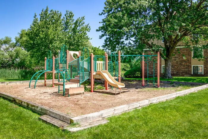 Apartments for Rent in Glendale Heights, IL - Ellyn Crossing Apartments - Outdoor Playground With Slide, Climbing Bars, Swing Set, and Mulch Area Beside Trees.