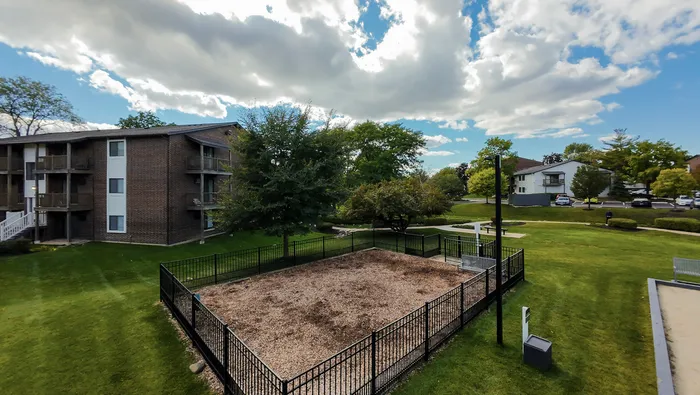 Palatine, IL, Apartments Near Roosevelt University - The Clayson - A Fenced Bark Park With Wood Chip Ground Cover In The Middle Of A Grass Lawn With Nearby Apartment Buildings.