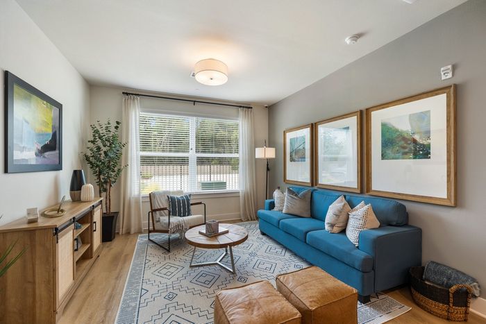 Houston, TX, Apartments with In-Unit Washer and Dryer - Acova Brentwood - Bright Living Room With Blue Sofa, Large Window, Wood-Style Plank Flooring, Framed Wall Art, And Modern Lighting.