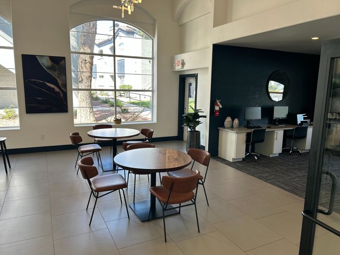 Modern communal area with stylish round tables and chairs, large arched windows for natural light, and a workspace featuring computers for residents.