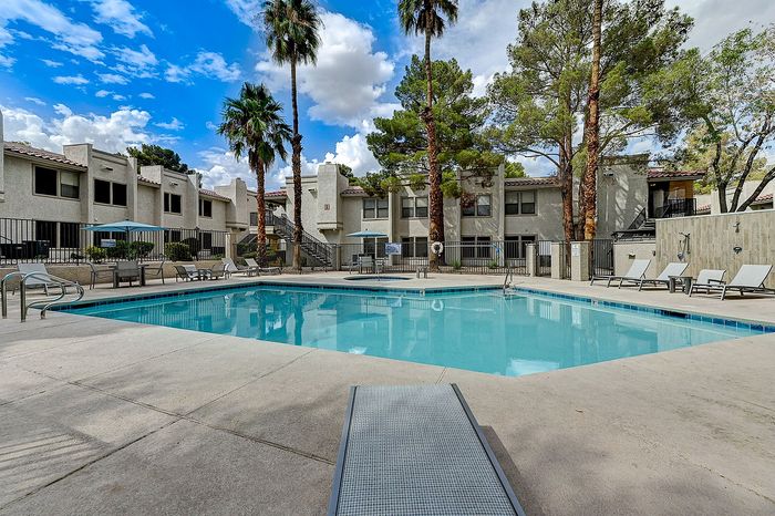 Inviting apartment complex with a sparkling swimming pool, surrounded by lush palm trees and stylish loungers, under a bright blue sky.