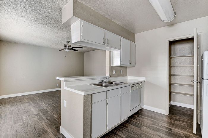 Modern kitchen with sleek white cabinetry, spacious countertops, and a convenient pantry door open, accented by elegant wood flooring and a ceiling fan in the adjacent living area.