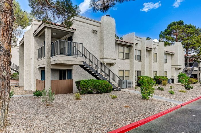 Charming two-story apartment building with modern architecture, nestled in a landscaped setting with mature trees and clear blue skies above.