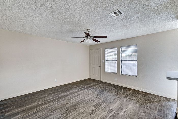Inviting living area with chic wood flooring, natural light streaming through expansive windows, and a modern ceiling fan offering comfort and style.