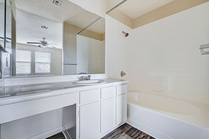 Bright and inviting bathroom featuring a sleek white vanity with ample storage, a large mirror, and a pristine bathtub, enhanced by natural light from nearby windows.