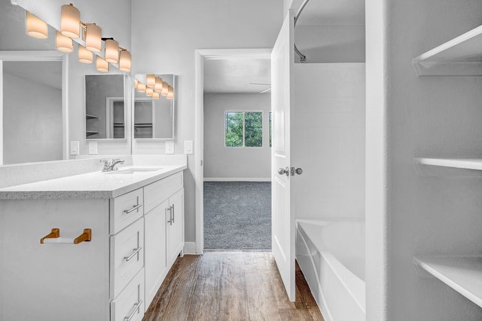 Inviting bathroom with elegant lighting, modern vanity, and sleek finishes, leading to a bright, carpeted bedroom with a scenic window view.