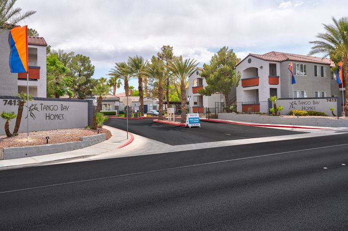 Inviting entrance to Tango Bay Homes featuring modern architecture, palm trees, and vibrant flags against a sunny backdrop.