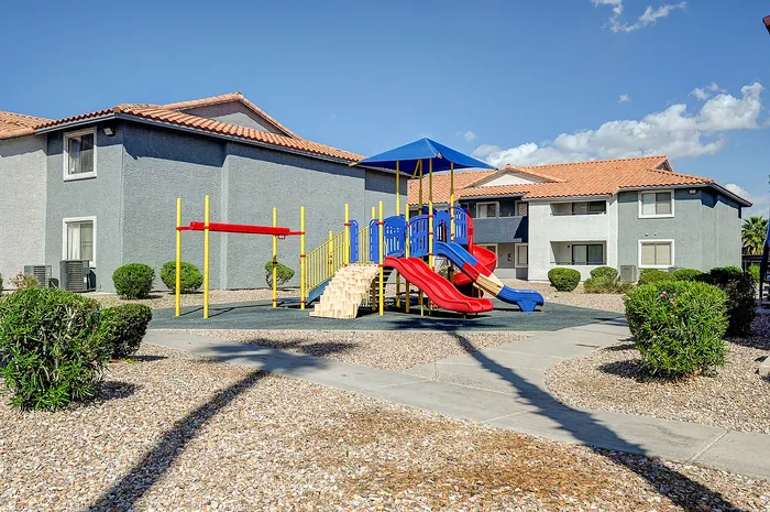 Charming apartment complex with a vibrant playground and beautiful landscaping under a sunny blue sky.