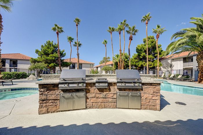 Luxury outdoor oasis featuring a sparkling pool, state-of-the-art stone barbecue area, and lush palm trees under a bright blue sky.