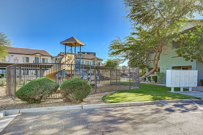 Vibrant apartment complex exterior with lush trees, a modern playground, and organized mailboxes under clear blue skies.