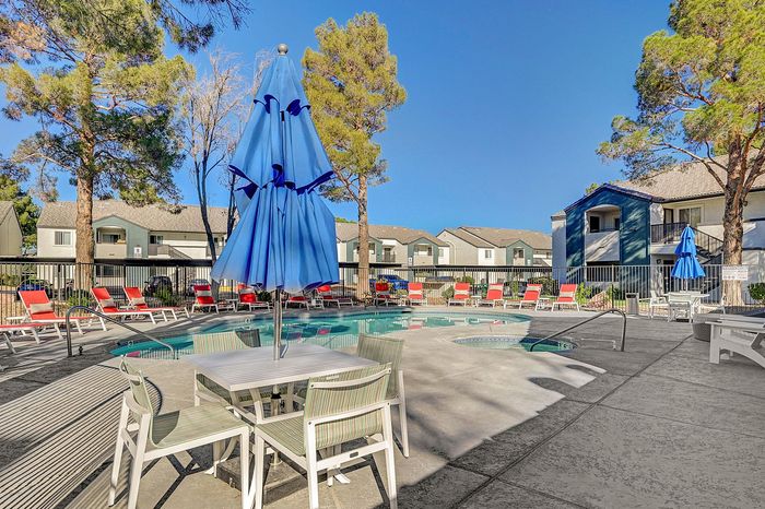 Inviting apartment complex pool area with vibrant red lounge chairs, blue umbrellas, and lush pine tree surroundings.
