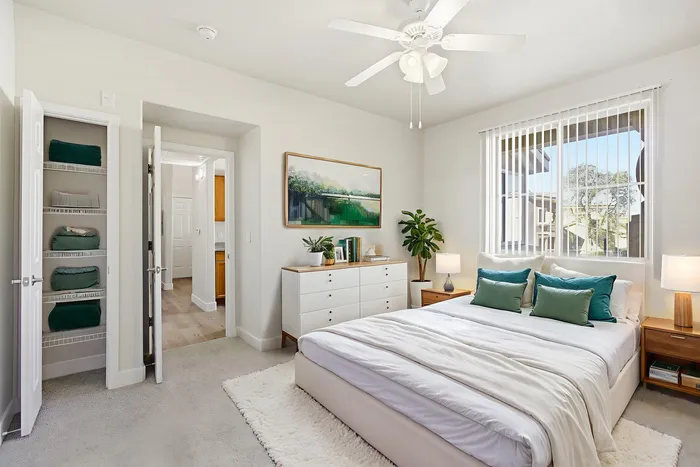 Bright and inviting bedroom with a plush bed, elegant decor, and ample natural light from large windows. Cozy and modern appeal with soft textures and greenery complementing the serene atmosphere.