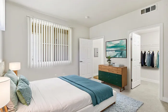 Bright and elegant bedroom showcasing a cozy bed with teal accents, stylish dresser, modern artwork, and a spacious walk-in closet.