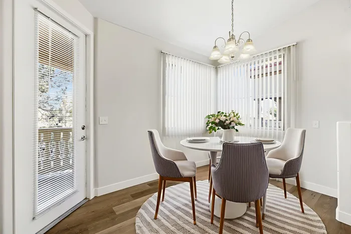 Bright dining area featuring elegant round table with chic chairs, large windows with vertical blinds, and a charming flower arrangement under a stylish chandelier.