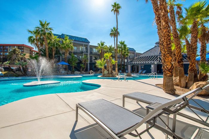 Luxurious apartment complex pool area with sparkling fountains, modern sun loungers, and towering palm trees under a clear blue sky.