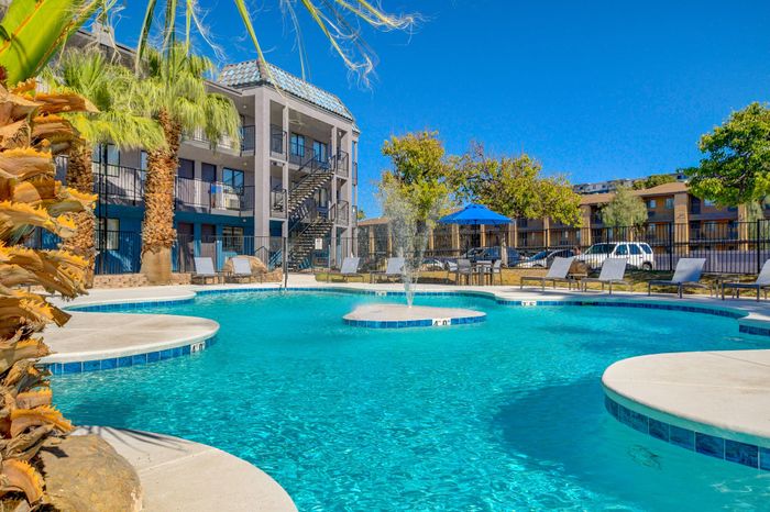 Luxurious apartment complex with a sparkling pool, sun loungers, and tropical palm trees under a clear blue sky.