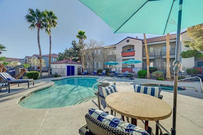 Charming apartment complex featuring a sunlit swimming pool, surrounded by palm trees, with stylish lounge chairs and inviting umbrellas for a perfect summer retreat.