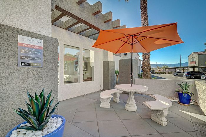 Inviting courtyard with elegant white benches and a vibrant orange umbrella, offering a chic outdoor seating area. Enhanced by lush green plants in modern blue pots, perfect for a relaxing atmosphere.