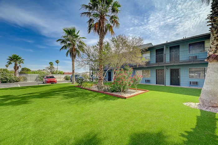 Charming apartment exterior with lush green lawn, vibrant landscaping, and towering palm trees under a clear blue sky.