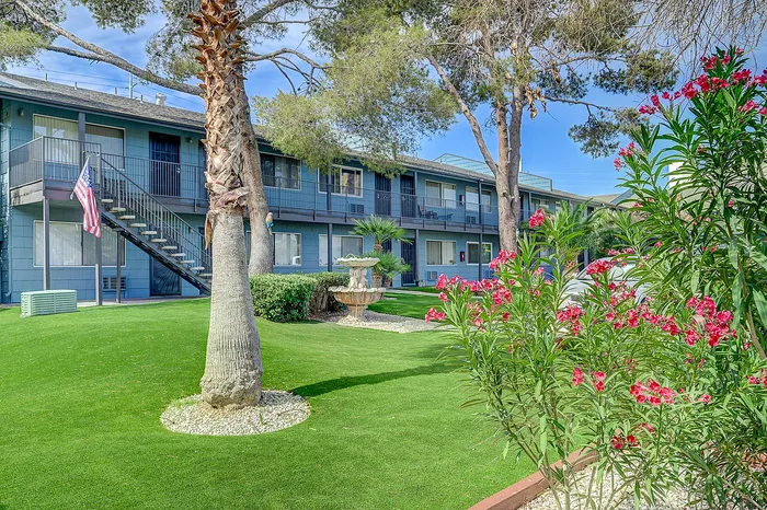 Charming apartment complex with lush green lawns, vibrant flowers, and a classic water fountain set among mature trees.