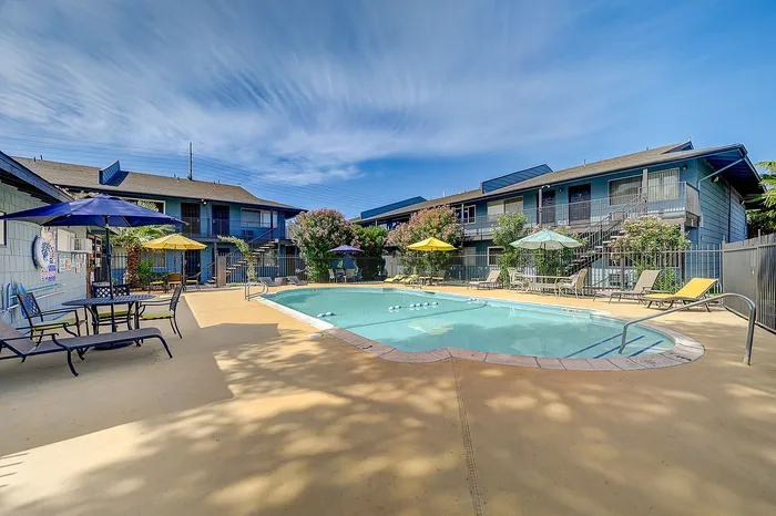 Charming apartment complex with a sparkling outdoor pool, surrounded by vibrant greenery and shaded seating areas under colorful umbrellas.