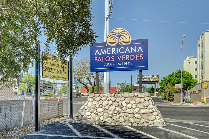 Inviting entrance to Americana Palos Verdes Apartments with vibrant signage and lush surroundings.