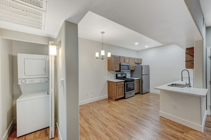 Modern kitchen and laundry area featuring sleek stainless steel appliances, elegant wood cabinetry, and a convenient stacked washer-dryer unit. Bright lighting enhances the open space with stylish hardwood flooring.